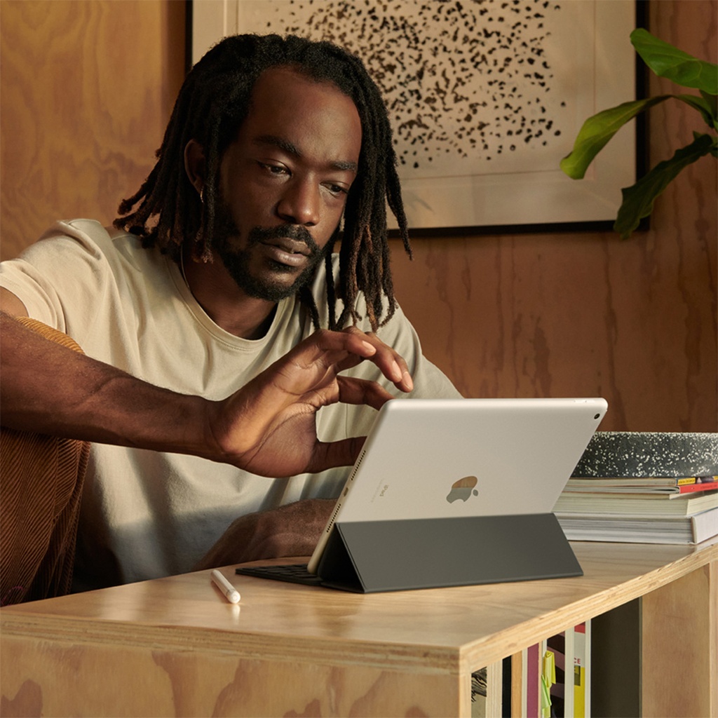 Un hombre con rastas y barba, vestido con una camiseta clara, está sentado frente a un iPad de 10.2 pulgadas de color plata, que está apoyado en un Smart Keyboard negro sobre una mesa de madera. El hombre mira atentamente la pantalla y extiende su mano derecha hacia ella. Hay un Apple Pencil blanco cerca del iPad y una pila de libros a la derecha.