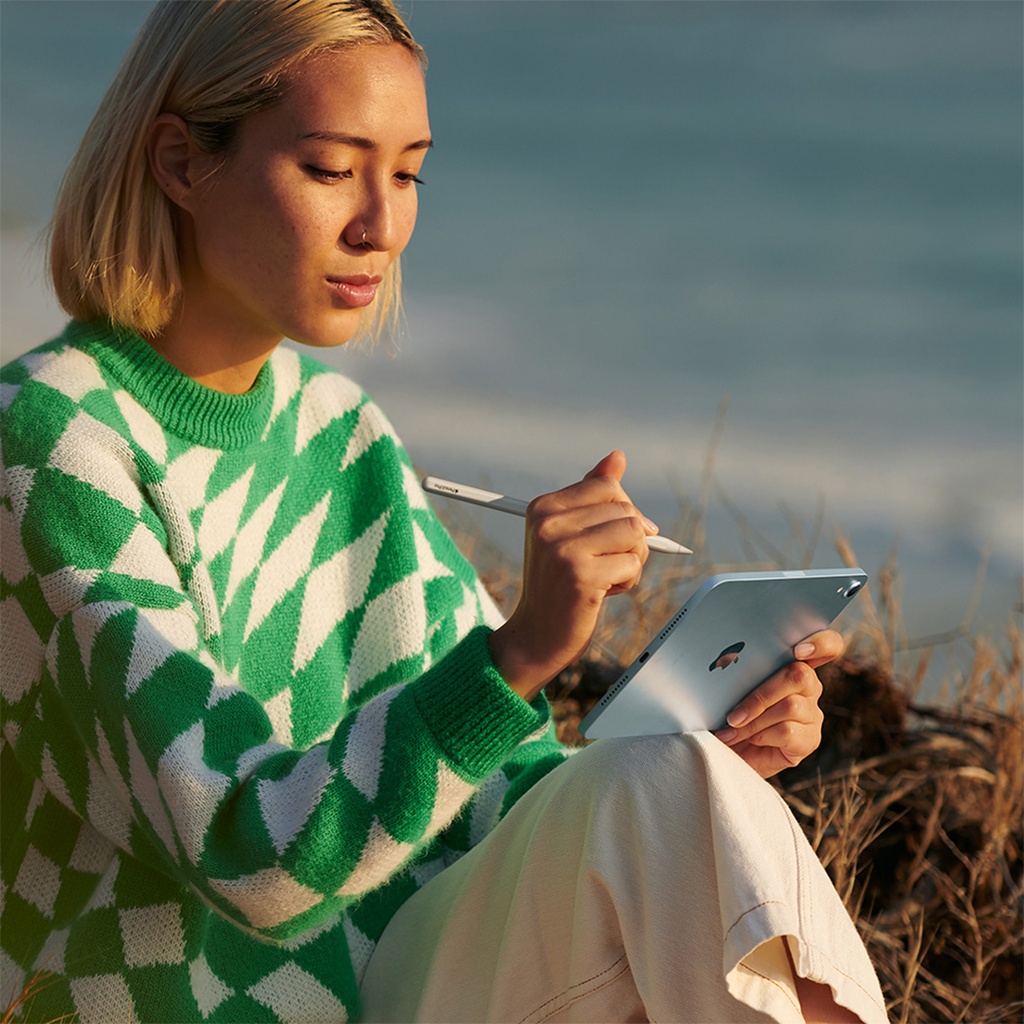 Una mujer joven de cabello corto y rubio, sentada al aire libre sobre la hierba seca, sostiene un iPad mini de color azul verdoso con la mano izquierda y usa un Apple Pencil blanco con la mano derecha sobre la pantalla. Viste un suéter verde y blanco con diseño de rombos. Al fondo se ve el mar o un lago, y el cielo despejado.