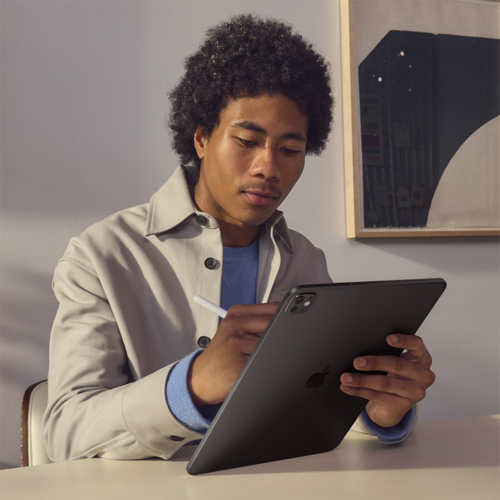 Un joven con cabello rizado y tez oscura, vestido con una camisa color crema y un suéter azul, está sentado en una silla moderna junto a una mesa. Sostiene un iPad Pro de 13 pulgadas de color negro espacial con su mano izquierda, y con su mano derecha usa un Apple Pencil blanco sobre la pantalla. El hombre mira atentamente el iPad, concentrado en la tarea.